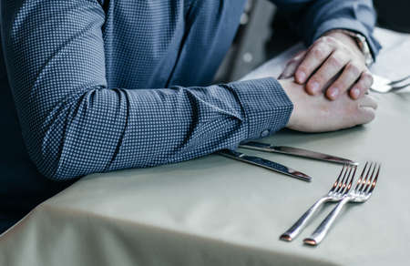 A young man in a blue shirt is sitting in a cafe and waiting for breakfast. Male hands next to the cutlery on the table.の写真素材
