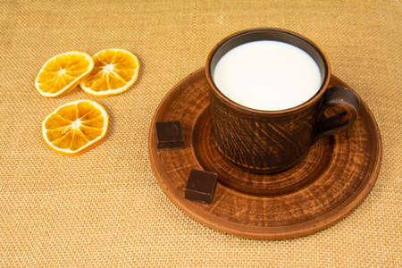 A mug of milk, two pieces of chocolate on a plate and orange slices on a table covered with burlap. Pottery on the table.の写真素材