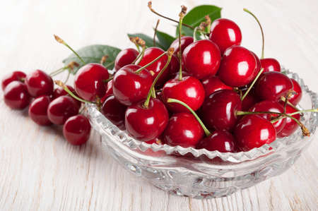 Red sweet cherry and green leaves in crystal vase on a wooden backgroundの写真素材
