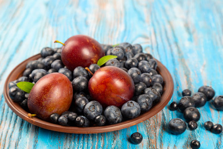 Fresh blueberries and plums in a brown plate on a blue wooden backgroundの写真素材