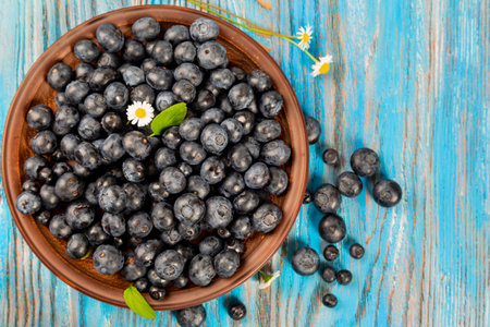 Fresh blueberries in a brown plate on a blue wooden background. Berries and daisy flowers, top viewの写真素材