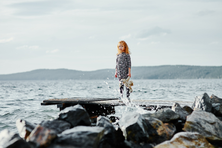 A sad child with a toy stands on the pier by the sea and looks at the splashes of waterの写真素材