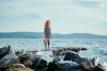 A sad child with a toy stands on the pier by the sea and looks at the splashes of waterの写真素材