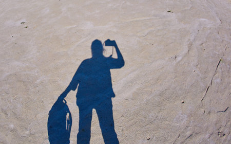 A self-portrait of a photographer with a camera in his hands casts a shadow on the sandy ground with...