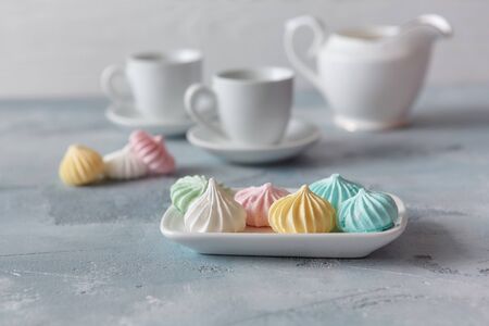 Multi-colored meringues in pastel shades with cups and a jug on a light textured wooden background. The concept of Breakfast with tea or coffee, home comfort and tenderness. Close-up, selective focus.の写真素材