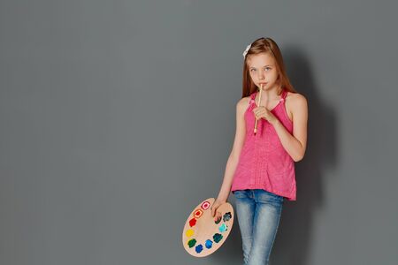 A pretty little girl with a palette and brushes against a gray wall ponders her future masterpiece.の写真素材