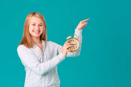 A teenage girl holds an alarm clock, standing on an isolated turquoise background, happy and laughing. Makes a hand gesture to an empty space for your text.の写真素材