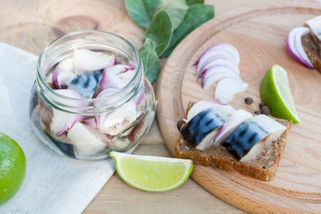 Slices of marinated mackerel with onion in a jar, lime, laurel and bread on wooden board, selective focusの写真素材