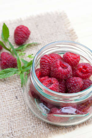 Fresh sweet red raspberry in a glass jar and mint on light wooden table, selective focusの写真素材
