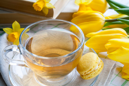 A cup of hot tea, yellow tulips, yellow daffodils, old books and lemon macaroons on a light...