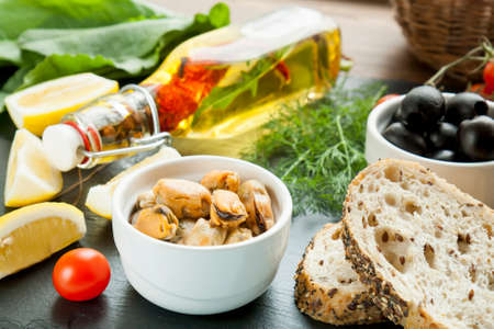Mussels and black olives and in ceramic bowls, fresh tomatoes, sliced lemon, bread and bottle of olive oil with spices on black stone board. Mediterranean food on black stone boardの写真素材