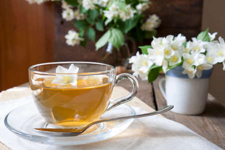 Transparent glass cup of tea with jasmine and blooming jasmine branch in blue ceramic cupon wooden boardの写真素材