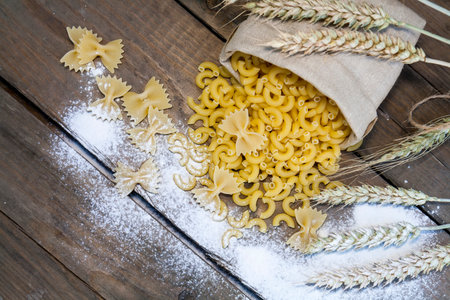 Uncooked Italian pasta Farfalle and Elbow macaroni in canvas little bag, scattered flour with wheat ears on dark wooden background.の写真素材