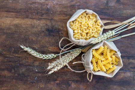 Uncooked Italian pasta Elbow macaroni and Fusilli in canvas little bag with wheat ears on dark wooden background. Shallow depth of fieldの写真素材