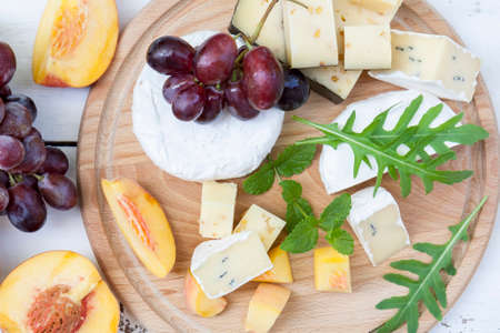 Different delicious cheeses camembert, blue cheese, cheese with nut, fruits red grapes and peach and herbs mint and arugula on wooden round boardの写真素材