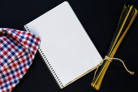 Blank cookbook with conventional and black uncooked Italian pasta Linguine, checkered napkin on...