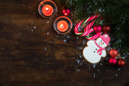 Christmas and New Year composition. Gingerbread snowman, fir tree branches, balls, light two candles and candy cane with snow on dark wooden background.の写真素材
