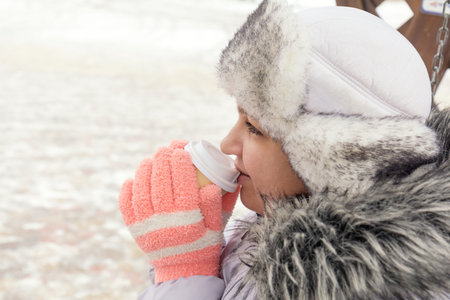 Young woman is drinking coffee on the street while walking on cold winter day. Hands with white take...