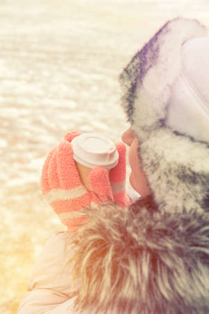 Young woman is drinking coffee on the street while walking on cold winter day. Hands with white take take-out coffee paper cup. Coloring and processing photo with soft focusの写真素材