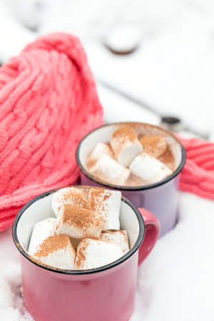 Hot chocolate with marshmallow in pink and violet two cups wrapped in a cozy winter pink scarf on the snow-covered table in the garden. Coloring and processing photo, small depth of fieldの写真素材