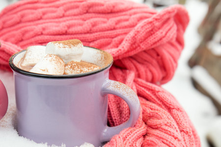 Hot chocolate with marshmallow in violet cup wrapped in a cozy winter pink scarf on the snow-covered table in the garden. Coloring and processing photo, selective focus, small depth of fieldの写真素材