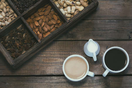 Cup of black coffee, cup of coffee with milk, cream. Almond, pistachios, anise, peanut and coffee beans in box. Dark wooden background. Beautiful vintage coffee groundwork. Coloring and processing photo.の写真素材