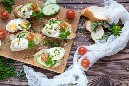 Bread with slices of fresh cucumber, egg, tomato and cream cheese on a wooden cutting board. Fresh parsley and rosemary.の写真素材