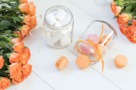 Orange mango or citrous macaroons and marshmallow in jars. Fresh little roses. Light wooden background. Sunlight. Coloring and processing photo with light vintage style. Shallow depth of field.の写真素材