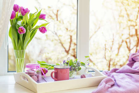 Cozy home concept. Purple fresh tulips in glass vase. Macaroons in glass jar. Cup of hot tea. White tray. Lilac blanket on the windowsill. Sunshine. Coloring and processing photoの写真素材