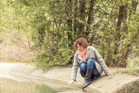 Pretty romantic young red head woman sitting close to the pond in spring park. Spring trees background.の写真素材