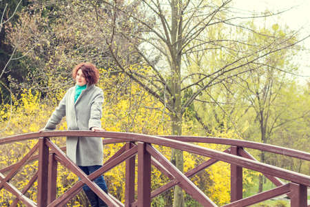 Pretty young red head woman on the bridge in spring park. Green trees and forsythia blossoms background.の写真素材