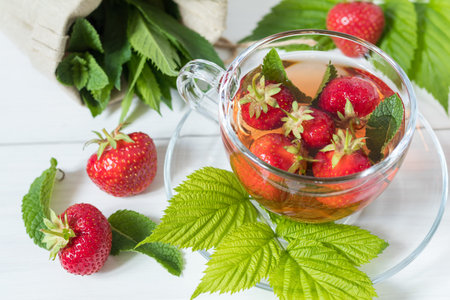 Glass cup of summer tea with fresh strawberry. Green leaves. Fresh mint. White wooden table. Shallow...