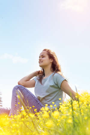 Young girl teenager on a lawn field with yellow flowers. Long chestnut hair. Sunshine, springtime, blue sky. Coloring and processing photos with soft selective focus. Shallow depth of field.の写真素材