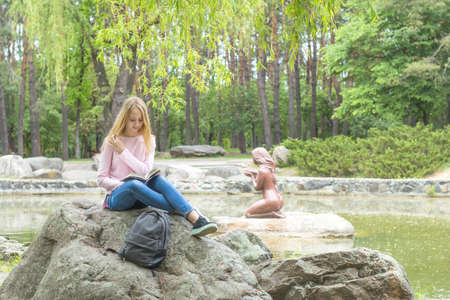 Cute romantic blond teenage-girl is reading a book sitting under the tree in the city park in the sunny weather. A pond surrounded by rocks with trees reflexion in the water is at the background.の写真素材