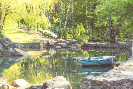 Sunny spring day in the city park with a pond surrounded by rocks. Trees reflection in the water. Blue wooden boat with two paddles peacefully resting in the pond.の写真素材