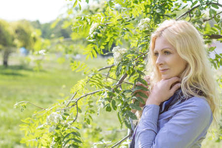 Pretty romantic blonde woman in the city park on a spring sunny day. Spring blossom and green leaves are at the background. Peaceful, quiet romantic atmosphereの写真素材