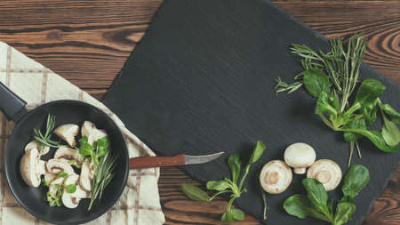 Top view of fresh mushrooms with mash salad and rosemary on a dark frying pan setting on a napkin near black stoned board on a wooden table. Copy spaceの写真素材