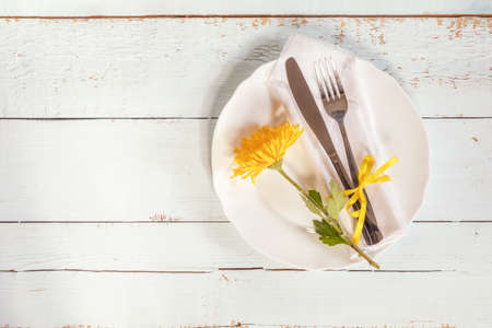 White empty plate, yellow chrysanthemum flowers, napkin, fork and knife tied with a yellow ribbon on light wooden background.の写真素材