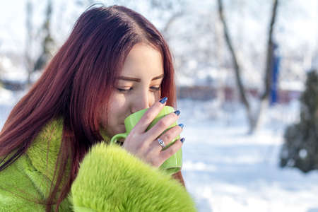 Beautiful cute pretty redhead female teenager in a green fur coat  drinking coffee in the sunny winter city park.の写真素材