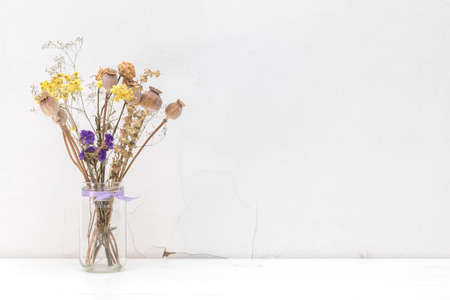 Composition of dried flowers and herbs, poppy heads in a glass jar with violet ribbon. White wooden surface close to the white cracked wall. Light tones, copy spaceの写真素材