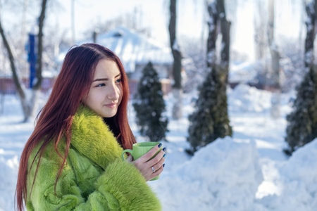 Beautiful cute pretty redhead female teenager in a green fur coat drinking coffee in the sunny winter city park.の写真素材