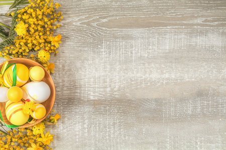Composition with painted eggs in a wooden bowl. Easter accessories, mimosa and yellow daffodils on a light wooden surface. Yellow orange easter concept, top view, copy space,  in yellow tones on a light wooden surfaceの写真素材