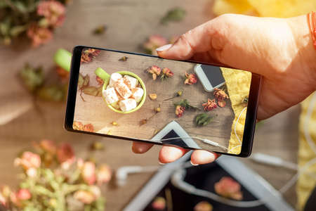 Woman hands taking food photo by mobile phone. Cup of coffee with marshmallow, white knitting wool, dried roses flowers, mobile phone and headphones. Beautiful romantic femaleâs background.の写真素材