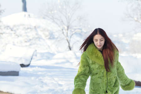 Beautiful cute pretty redhead female teenager in a green fur coat in the sunny winter city park.の写真素材
