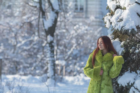 Beautiful cute pretty redhead female teenager in a green fur coat in the sunny winter city park.の写真素材