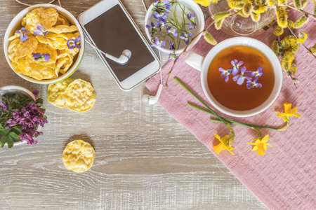 Cup of tea with violet viola, mobile phone and headphones, delicious nutritious cereal breads and spring flowers on wooden table. Delicious nutritious breakfast, top view, copy space.の写真素材