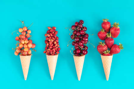 Four waffle cones with red cherries, wild strawberries, yellow cherries, and strawberryes on blue surface. Flat lay, top view sweet food background.の写真素材