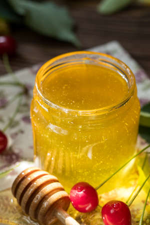 Open glass jar of liquid honey and honey dipper, bunch of linden flowers and red cherry on wooden surface. Ray of sunlight. Dark rustic style.の写真素材