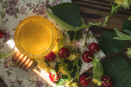 Open glass jar of liquid honey and honey dipper, bunch of linden flowers and red cherry on wooden...