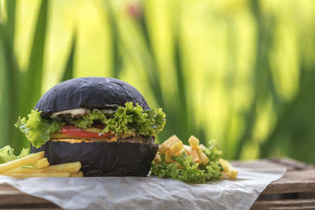 Burger made with black charcoal bun served with potato wedges, lettuce and sauce on wooden rustic table Close up, shallow depth of the field.の写真素材